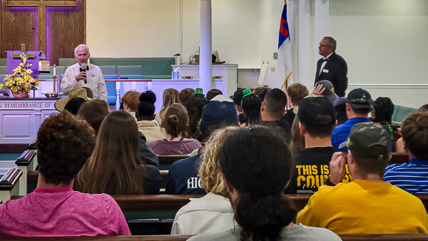 Man with microphone speaks to young crowd in sanctuary.