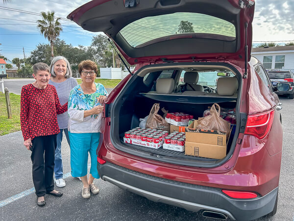 Three women stand beside car loaded with soup for charity.