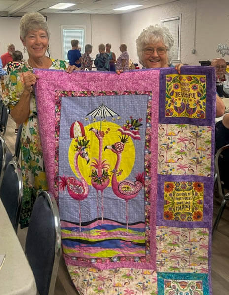 Two ladies hold up a quilt with flamingos and inspirational sayings.
